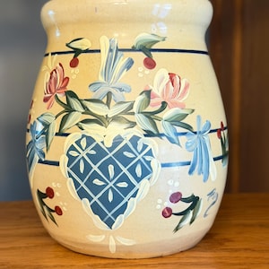 May include: A cream-colored ceramic crock featuring hand-painted floral motifs in shades of blue, pink, and green. The design includes a central blue heart with a white lattice pattern, accented by horizontal blue stripes and small red berries.