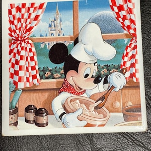 May include: Square ceramic tile featuring Mickey Mouse in a chef's hat, stirring a bowl. The background shows a window with red and white checkered curtains, a castle, and a dome. The tile has a glossy finish and the Disney logo.