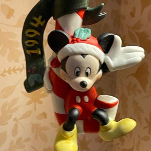 May include: A Mickey Mouse Christmas ornament from 1994. Mickey is wearing a Santa hat and red outfit, holding a white bucket. The ornament is attached to a candy cane with a green banner that reads "1994".