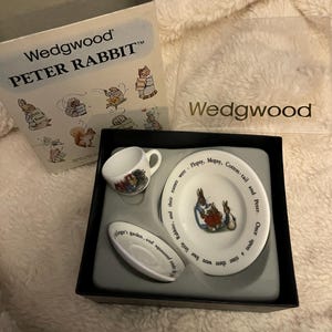May include: A boxed Wedgwood Peter Rabbit tea set. The set includes a small teacup, a saucer, and a plate. The plate and cup feature illustrations of Peter Rabbit and other characters. The box and the set are white.