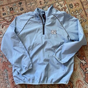K&ouml;nnte beinhalten: Eine hellblaue Windbreaker-Jacke mit Rei&szlig;verschluss und grauem Besatz. Die Jacke hat ein Logo auf der Vorderseite, das "Tar Heels" sagt und ein blaues und wei&szlig;es "N" mit wei&szlig;em Umriss zeigt.