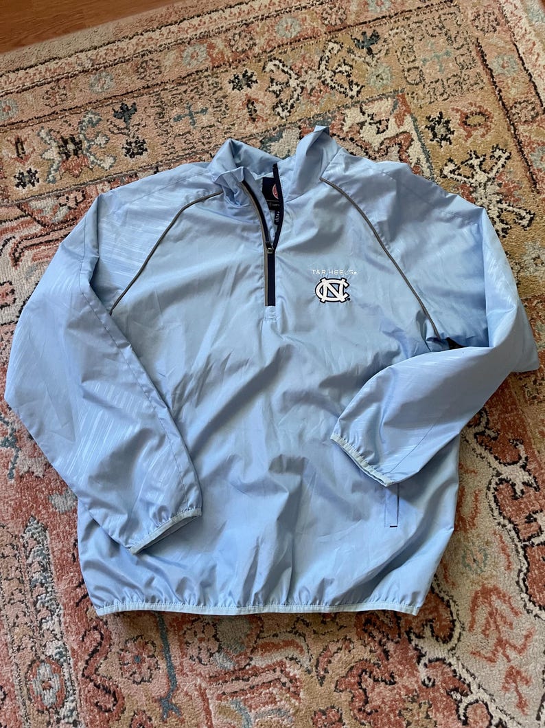 K&ouml;nnte beinhalten: Eine hellblaue Windbreaker-Jacke mit Rei&szlig;verschluss und grauem Besatz. Die Jacke hat ein Logo auf der Vorderseite, das "Tar Heels" sagt und ein blaues und wei&szlig;es "N" mit wei&szlig;em Umriss zeigt.