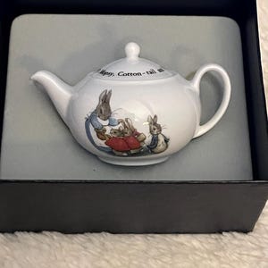 May include: White ceramic teapot with a lid, featuring a handle and spout. The teapot is decorated with illustrations of rabbits in clothing. The text "Mopsy, Cotton-tail" is printed on the teapot. The teapot is presented in a black box.