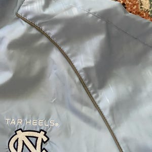 K&ouml;nnte beinhalten: Hellblaue Windbreaker-Jacke mit wei&szlig;em und grauem Besatz. Die Jacke tr&auml;gt die Worte "TAR HEELS" und ein blaues und wei&szlig;es "N"-Logo, das auf der Vorderseite gestickt ist.