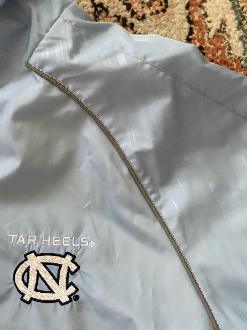 K&ouml;nnte beinhalten: Hellblaue Windbreaker-Jacke mit wei&szlig;em und grauem Besatz. Die Jacke tr&auml;gt die Worte "TAR HEELS" und ein blaues und wei&szlig;es "N"-Logo, das auf der Vorderseite gestickt ist.