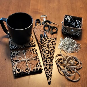 May include: A black mug sits on a leopard print coaster next to a matching set of coasters tied with twine. A leopard print scissor holder contains scissors and pens. A small box of binder clips and a pile of paper clips and rubber bands are also present.