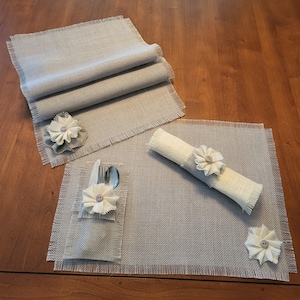May include: A rustic dining set featuring a gray burlap runner, placemats, a napkin ring, and a utensil holder. Each piece is adorned with a fabric flower and a button center, creating a cohesive and charming table setting.