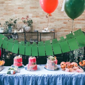 May include: A table with a blue tablecloth is set with three pink and white layered cakes, a bowl of fruit, and a green banner with the text "Happy Birthday". There are also two balloons, one red and one green.