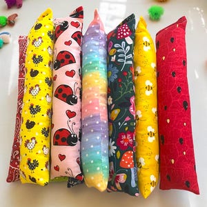 May include: Seven long, rectangular fabric pillows arranged side-by-side. Patterns include red paisley, yellow with black chickens, pink with ladybugs and hearts, a rainbow gradient with white dots, dark green with flowers, yellow with bees, and red with seeds.