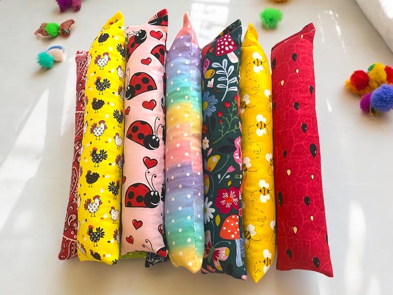 May include: Seven long, rectangular fabric pillows arranged side-by-side. Patterns include red paisley, yellow with black chickens, pink with ladybugs and hearts, a rainbow gradient with white dots, dark green with flowers, yellow with bees, and red with seeds.