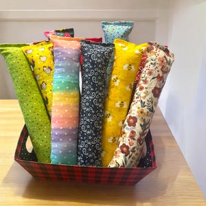 May include: A collection of long, cylindrical fabric pouches in a red and black plaid basket. The pouches feature various patterns including green with dots, yellow with chickens, rainbow stripes, navy blue paisley, yellow with bees, and floral prints.