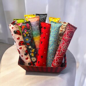 May include: A collection of long, cylindrical fabric pouches with various patterns including ladybugs, butterflies, rainbow dots, red with black seeds, blue with colorful circles, and red paisley, all standing upright in a red and black plaid basket.