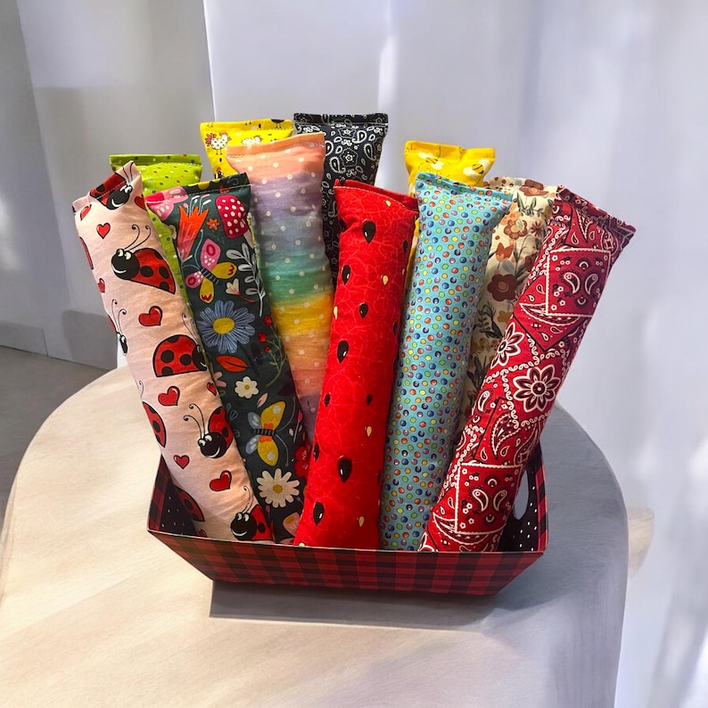 May include: A collection of long, cylindrical fabric pouches with various patterns including ladybugs, butterflies, rainbow dots, red with black seeds, blue with colorful circles, and red paisley, all standing upright in a red and black plaid basket.