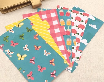 May include: A collection of colorful planner dividers with various patterns. Designs include butterflies, yellow stripes, pink gingham, juice drinks, red turtles, and white clouds on a blue background. A gold planner cover is visible in the background.
