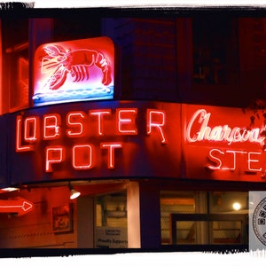 May include: A nighttime shot of a restaurant facade with neon signs. The sign reads "Lobster Pot" in large red letters, with a neon lobster graphic above. Additional neon signs advertise "Charcoal Broiled Steaks".