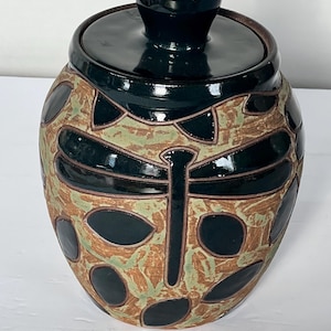 May include: A black ceramic jar with a lid and a dragonfly design. The jar has a brown and green speckled glaze with black accents.