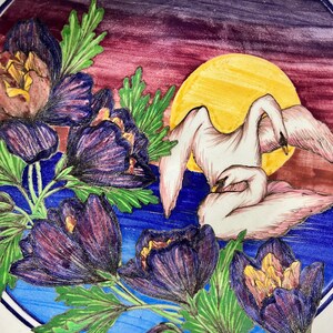 May include: A decorative plate with a hand-painted design. The plate features purple flowers, green leaves, and two white swans in front of a yellow sun. The background transitions from blue to purple.