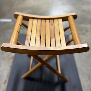 以下が含まれることがあります： スラットシートと湾曲した上部および下部レールを備えた、明るい茶色の折りたたみ式木製スツール。脚は安定性のために交差して角度が付けられています。スツールは、携帯性とコンパクトな収納のために設計されています。