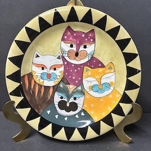 May include: A round ceramic plate with a yellow background and a black and white starburst border. Four cats with different colored fur and facial features are painted on the plate.
