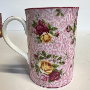 Puede incluir: Una taza alta de cerámica con asa blanca y diseño floral rosa. La taza presenta rosas amarillas, rojas y rosas sobre un fondo rosa con un sutil diseño de cachemira. El borde está adornado con un rosa más oscuro.