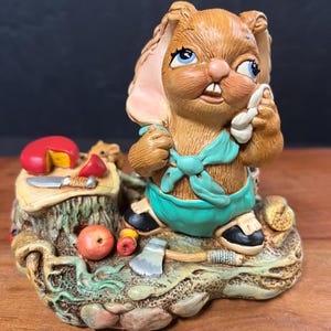 Peut inclure: Figurine fantaisiste d'un lapin brun aux yeux bleus et une écharpe turquoise, tenant un objet blanc. Le lapin est sur une base détaillée avec un morceau de fromage, une pomme et une petite hache. La figurine a un aspect rustique et peint à la main.