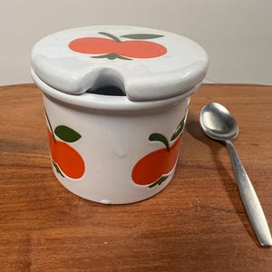May include: A white ceramic jar with a lid, adorned with orange and green apple designs. A small, silver spoon rests beside the jar. The jar is on a wooden surface.