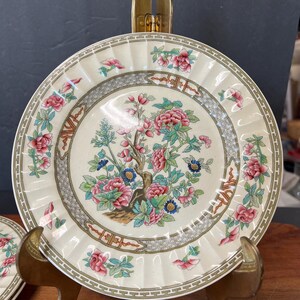 May include: Cream-colored ceramic plate with a floral design. The plate features a central image of a flowering tree with pink and blue blossoms. The rim has a repeating pattern and additional floral accents. The plate is displayed on a gold-colored stand.