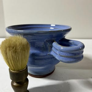 Op de afbeelding: Een blauwe keramische scheerkom met een handvat en een scheerkwast. De kom heeft een brede rand en een klein handvat. De scheerkwast heeft een donker handvat en lichte haren.