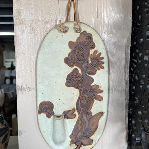 May include: A white ceramic wall vase with a brown floral design and a single opening for flowers. The vase has a leather strap for hanging.