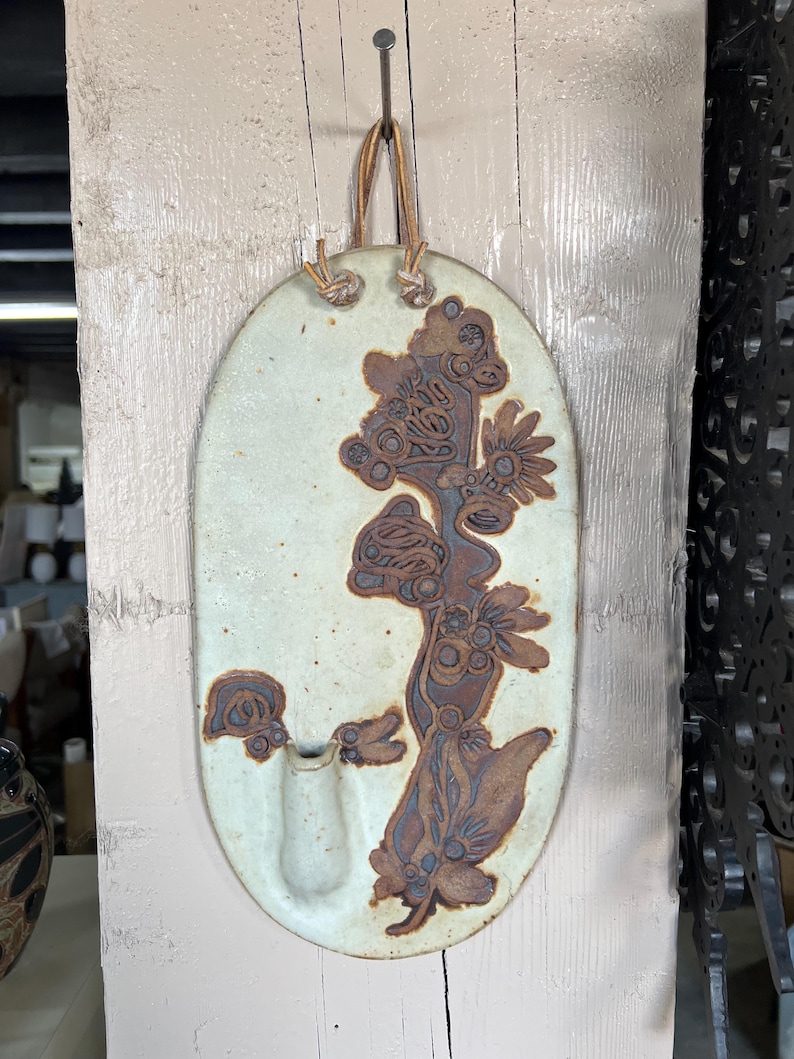 May include: A white ceramic wall vase with a brown floral design and a single opening for flowers. The vase has a leather strap for hanging.