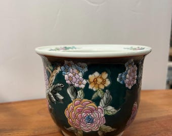 May include: A ceramic planter with a dark green background and floral design. The planter has a white rim and a detailed pattern of pink, yellow, and blue flowers with green leaves. The planter is on a wooden surface.