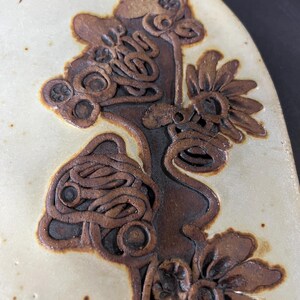 May include: A ceramic wall hanging featuring a brown floral design. The design incorporates stylised flowers with intricate details and a textured surface.