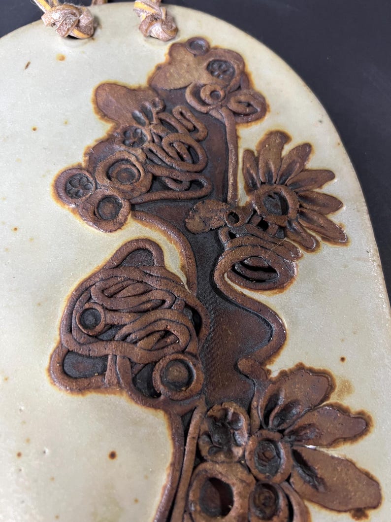 May include: A ceramic wall hanging featuring a brown floral design. The design incorporates stylised flowers with intricate details and a textured surface.