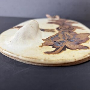 May include: A white ceramic wall hanging with a brown floral design. The hanging has a raised edge and a slightly curved shape.