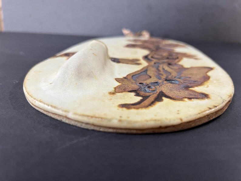 May include: A white ceramic wall hanging with a brown floral design. The hanging has a raised edge and a slightly curved shape.