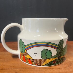 May include: White ceramic pitcher with a curved handle and spout. The pitcher features a colorful landscape design with a rainbow, trees, and fields. The design is in shades of green, yellow, orange, and red. The pitcher is sitting on a wooden surface.