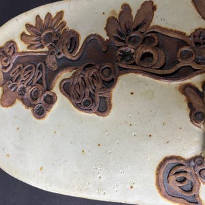 May include: A white ceramic plate with a brown floral design. The design features intricate swirls and loops, creating a unique and artistic pattern.
