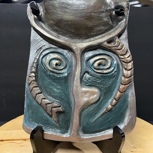 Puede incluir: Escultura de cerámica abstracta tallada a mano en tonos grises, azules y marrones. La obra presenta un rostro estilizado con ojos en espiral y detalles serpentinos. La escultura se exhibe sobre un soporte de madera negro.
