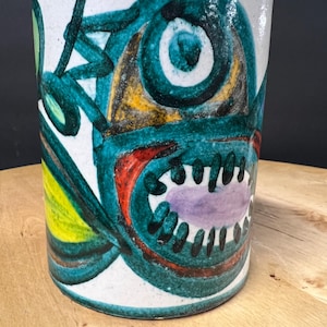 May include: A cylindrical ceramic mug with a hand-painted abstract design. The artwork features a teal, yellow, red, and purple creature-like face against a white background. The mug is approximately 5 inches tall.
