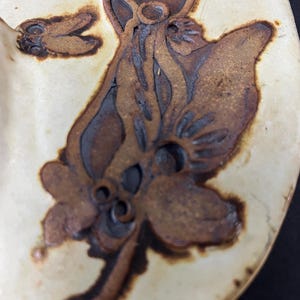 May include: A ceramic plate with a brown glaze features a stylised floral design. The design is embossed and has a raised texture. The plate is white with a brown rim.