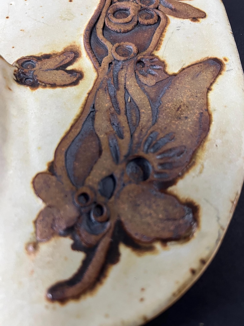 May include: A ceramic plate with a brown glaze features a stylised floral design. The design is embossed and has a raised texture. The plate is white with a brown rim.