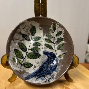 May include: A ceramic bowl with a brown exterior and a white and brown interior. The bowl features a blue bird design with green leaf accents. The bowl is displayed on a gold-colored metal stand.