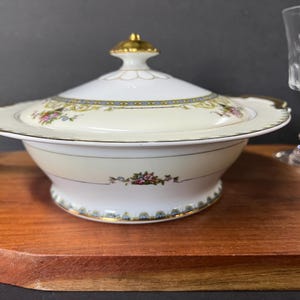 May include: Cream-coloured covered serving dish with gold trim and floral accents. The lid has a gold finial. The dish sits on a wooden surface. A clear glass is in the background.