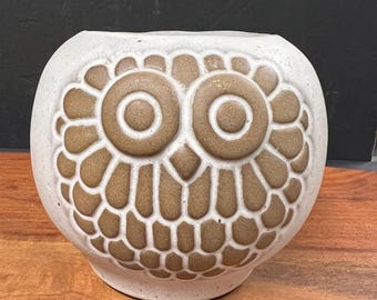 May include: A cream-coloured ceramic owl-shaped vase with brown detailing. The owl's face is the focal point, with large, round eyes and a textured pattern. The vase has a rounded shape and a wide opening, suitable for flowers or other decorative items.