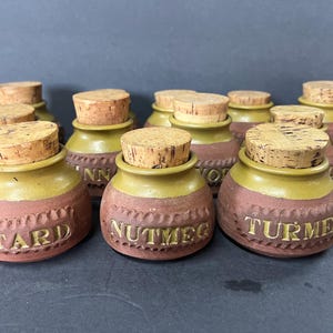 Puede incluir: Un juego de diez pequeños tarros de especias de cerámica marrón con tapas de corcho. Cada tarro tiene un nombre de especia diferente escrito en la parte delantera, incluyendo "Star", "Cinnamon", "Nutmeg", "Turmeric" y "Pepper".