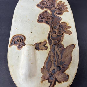 May include: A white ceramic wall hanging with a brown floral design. The hanging has a small, oval-shaped pocket on the left side.