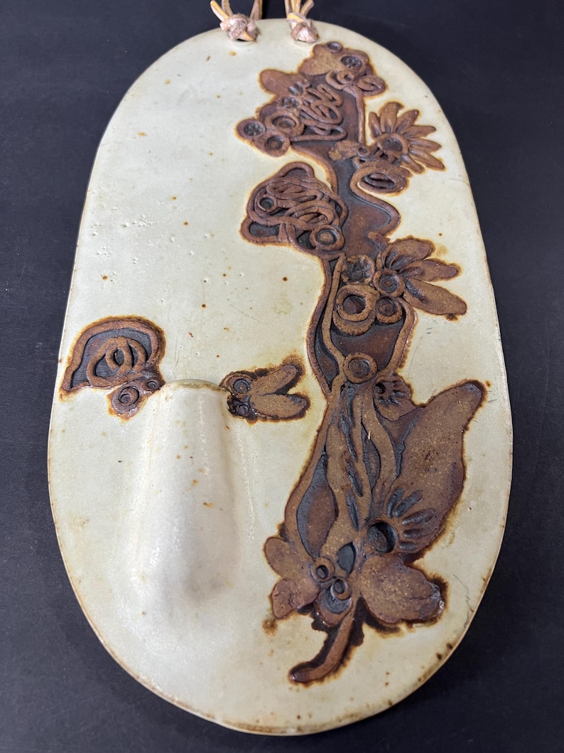May include: A white ceramic wall hanging with a brown floral design. The hanging has a small, oval-shaped pocket on the left side.