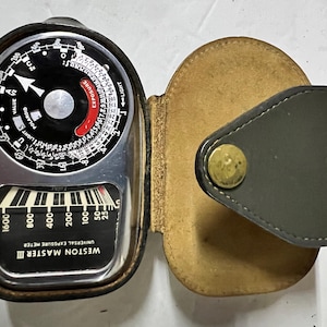 Puede incluir: Un fotómetro universal Weston Master II plateado con una esfera negra y una funda de cuero. El fotómetro tiene un rango de 25 a 1600.