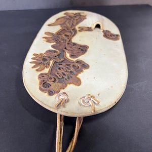 May include: A white ceramic wall hanging with a brown floral design and a hole at the top. The hanging has a leather strap with two knots.