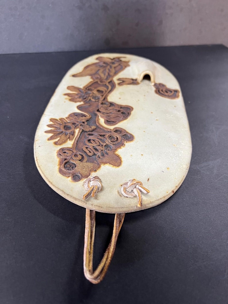 May include: A white ceramic wall hanging with a brown floral design and a hole at the top. The hanging has a leather strap with two knots.